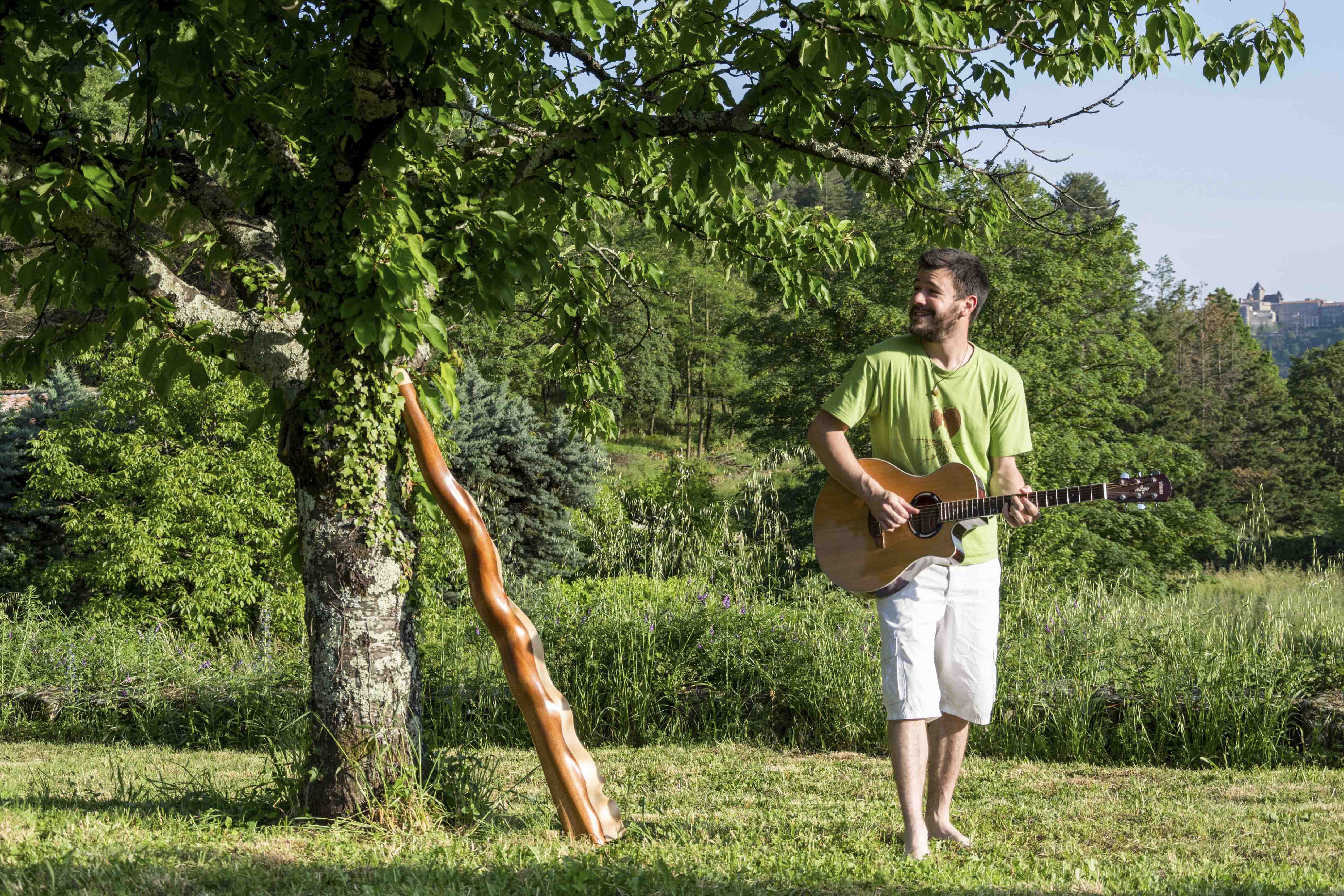 Photo de Julien Doutaz, fabricant des ujazi didgeridoos, au pied d'un arbre avec une guitare et un didgeridoo sculpté